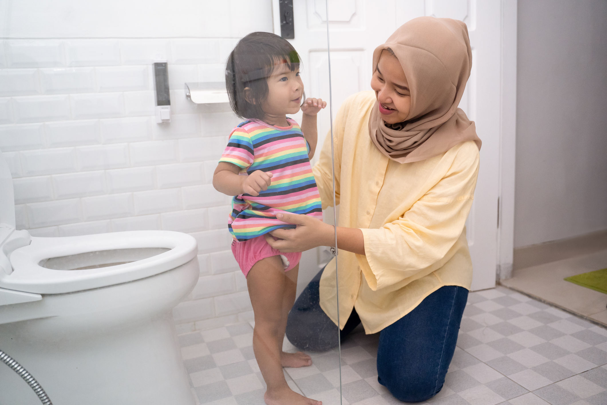 Toddler stands next to a toilet in a bathroom with parent while the parent fixes the toddler's clothes.