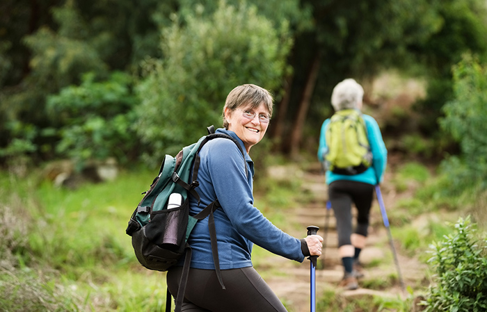 Two senior women hiking along path wearing backpacks and using hiking sticks