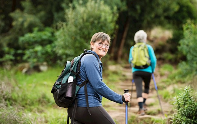 Two senior women hiking along path wearing backpacks and using hiking sticks