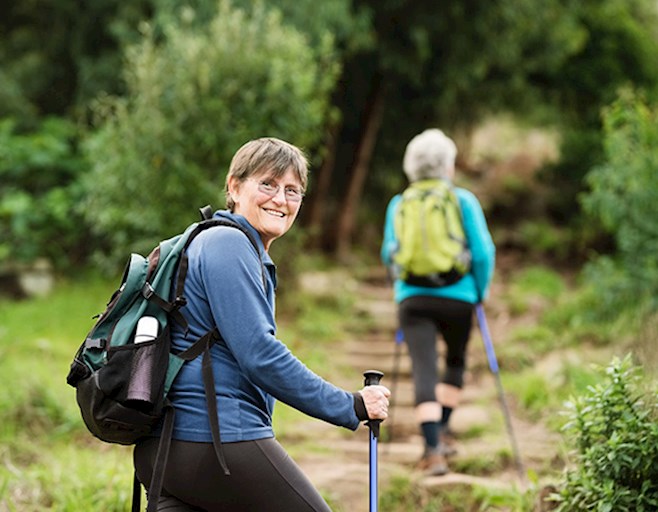 Two senior women hiking along path wearing backpacks and using hiking sticks
