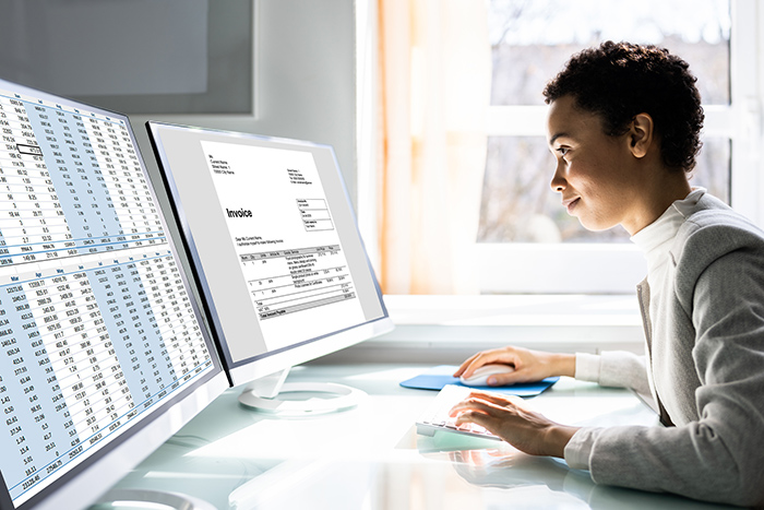 Woman sitting at desk looking at two large computer monitors looking at spreadsheets