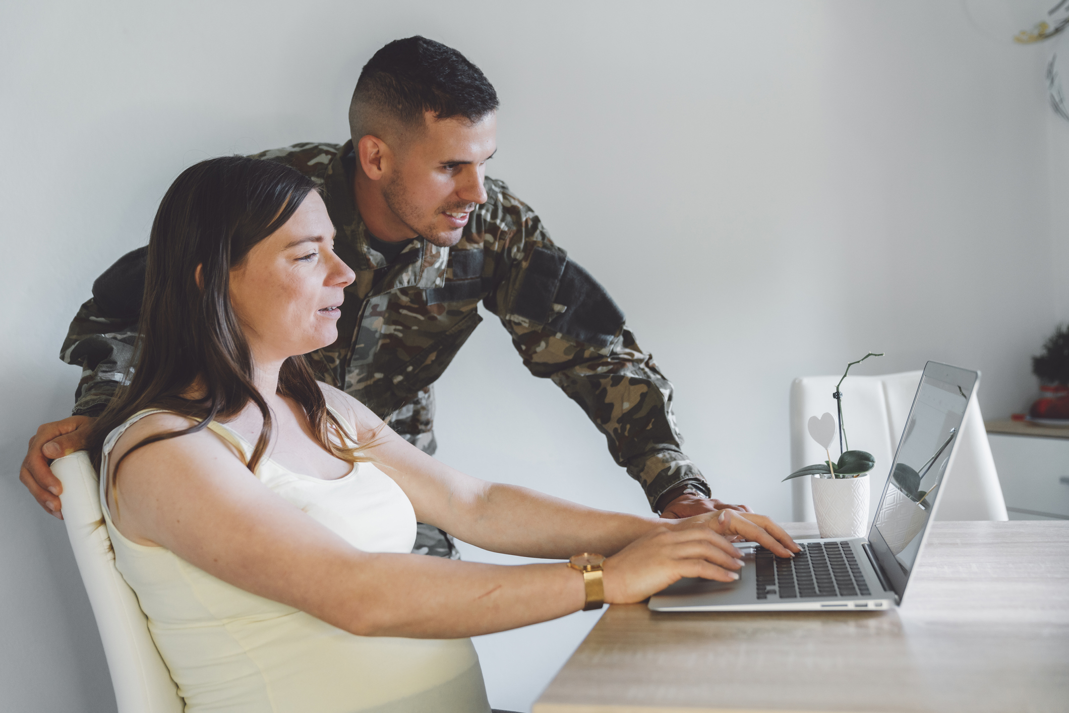 Woman sitting at desk on laptop. Man in military fatigues standing next to her looking at the laptop.