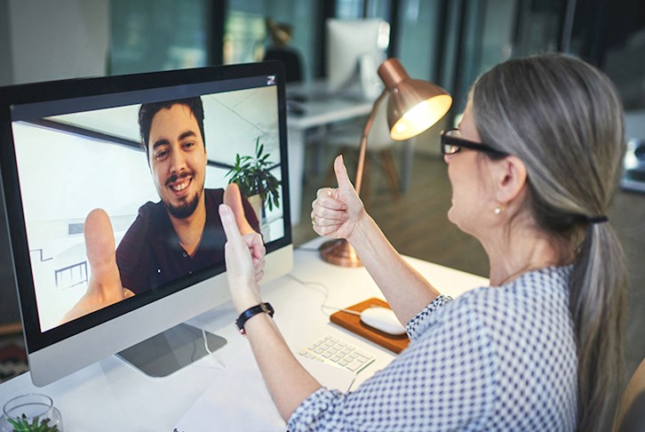 Woman sitting at desk giving thumbs up to man in computer monitor giving thumbs up during telehealth conference
