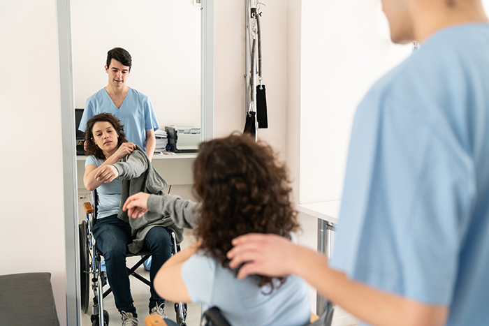 Woman in wheelchair looking in mirror while putting jacket on with healthcare worker behind her