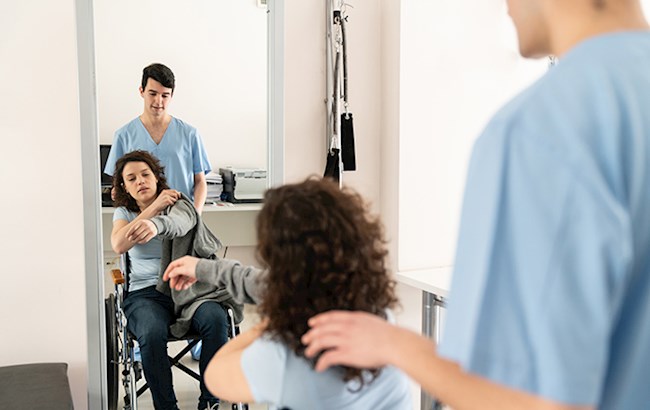 Woman in wheelchair looking in mirror while putting jacket on with healthcare worker behind her