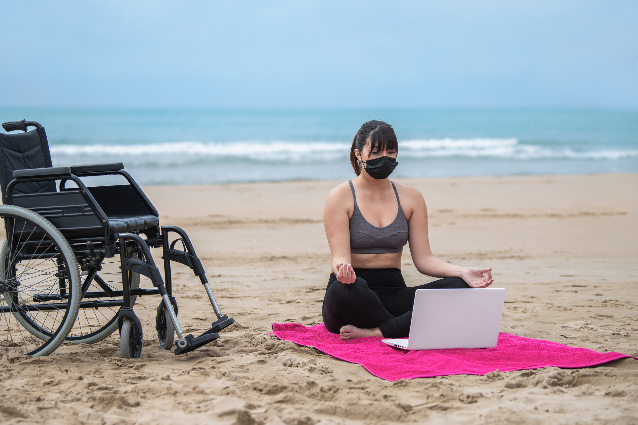Woman sitting cross-legged on beach with open laptop. Wheelchair is next to her on the sand.