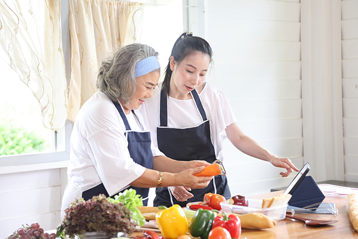 Two women wearing aprons cooking with fresh food and looking at tablet on table