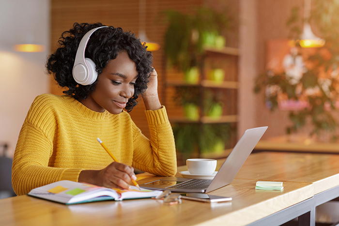 Woman sitting at table wearing headphones and working on laptop while writing in notebook
