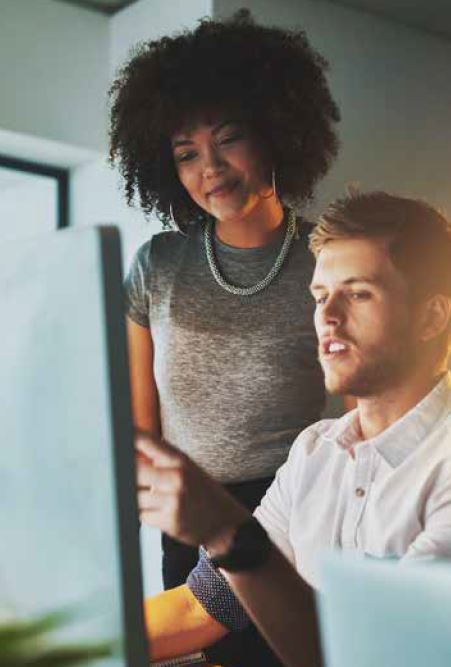 Woman smiling, standing by man who is sitting and using a laptop.