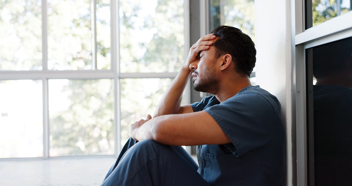 Man sits on floor with his hand on his forehead and other arm across his knee, staring out the window.
