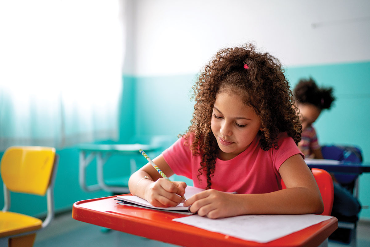 Young student writing at a red desk with another student in the background at a blue desk