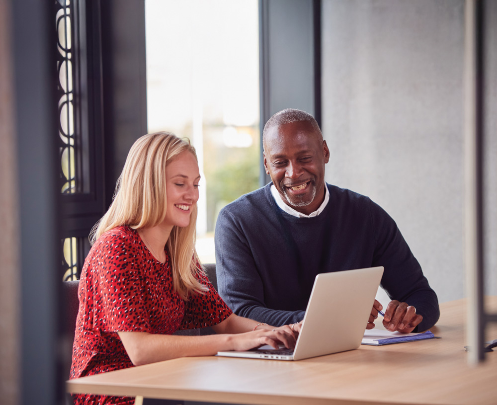 Woman and man, smiling, sitting at a desk and looking at a laptop.