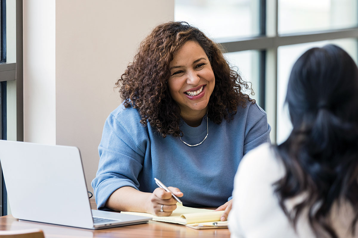 Woman with a laptop nearby, holding a pen and smiling at the woman across the table from her