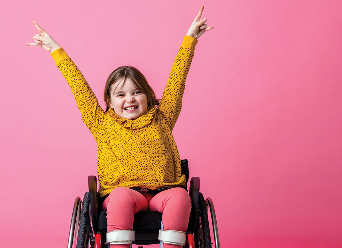 Young girl wearing a yellow top, sitting in a wheelchair, against a pink background