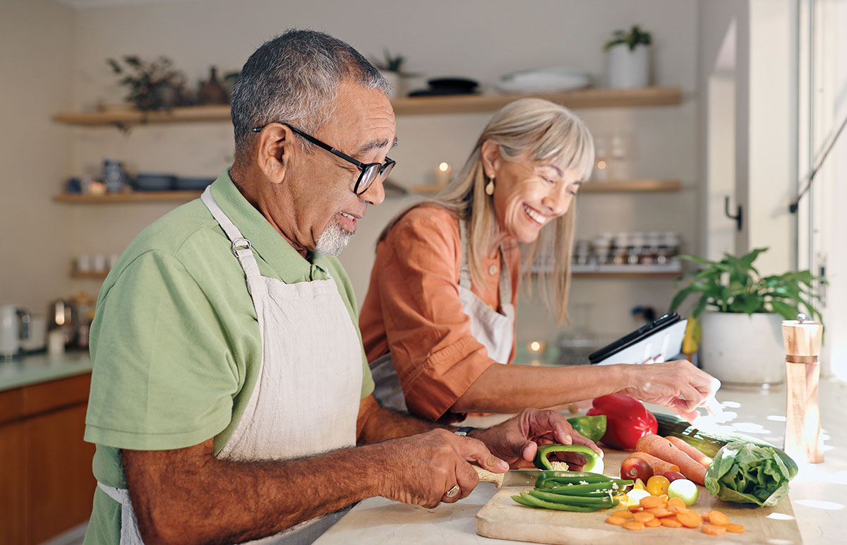 An older man with glasses and a goatee chopping vegetables with a smiling woman in a kitchen setting