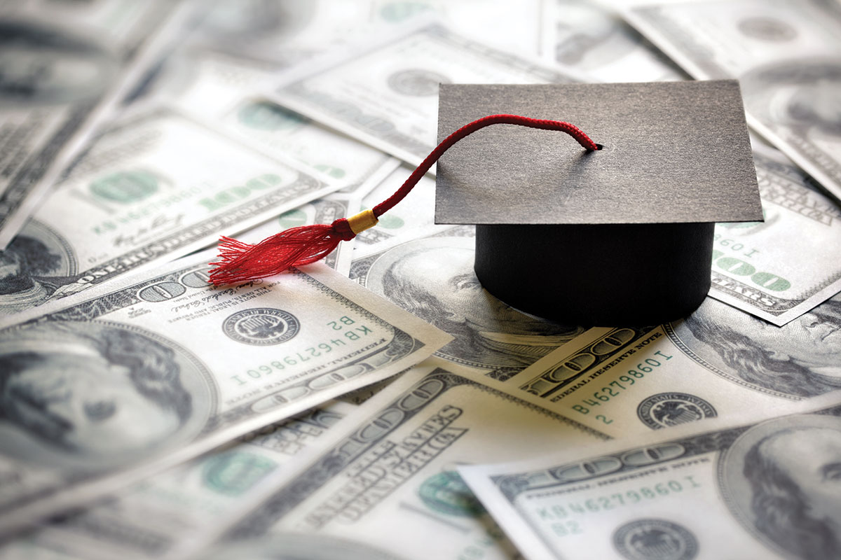 A stock photo of a mortar board with a red tassel sitting on a lot of $100 bills