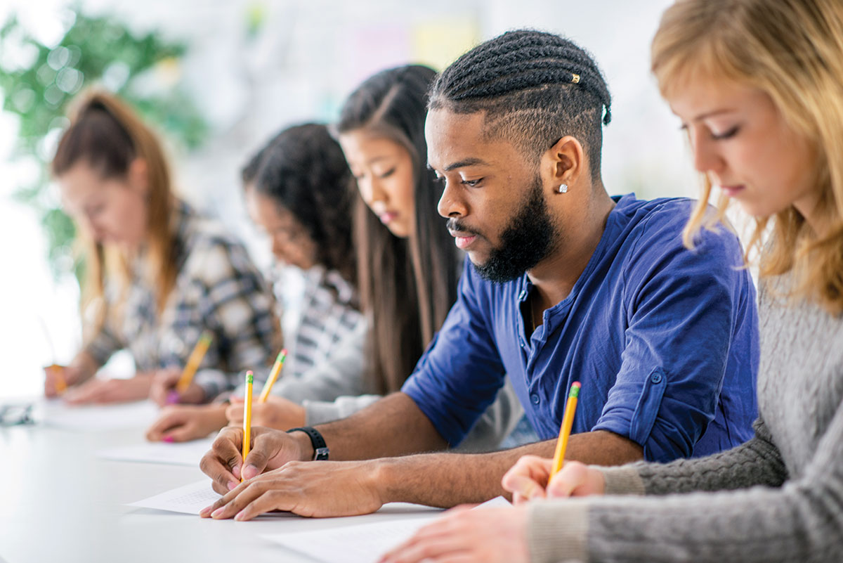 A group of young people sitting at a table writing quietly with their eyes looking down.