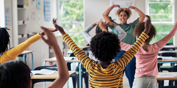 Teacher standing in front of young students, and all in the classroom are extending their arms and moving and smiling