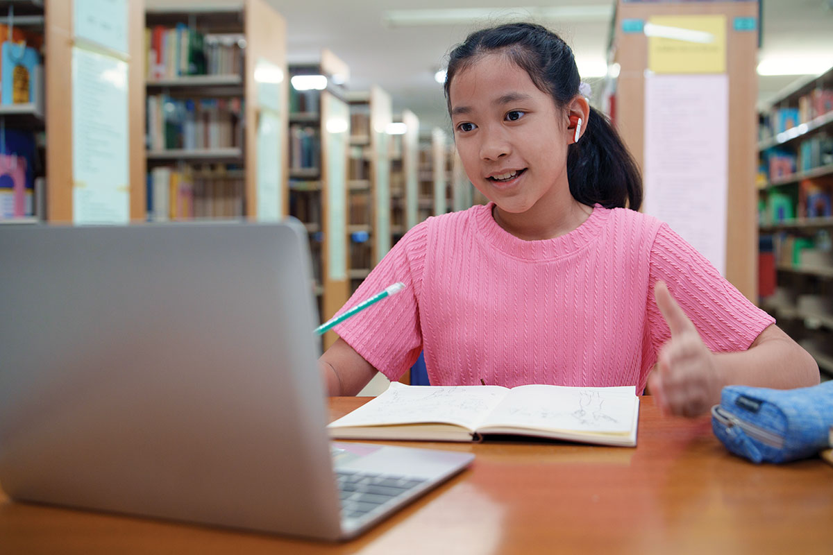 Young girl in library setting watching something on laptop, taking notes using pencil and paper
