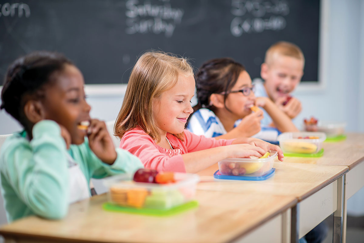 Young children sitting lunch at a table eating with a blackboard in the background