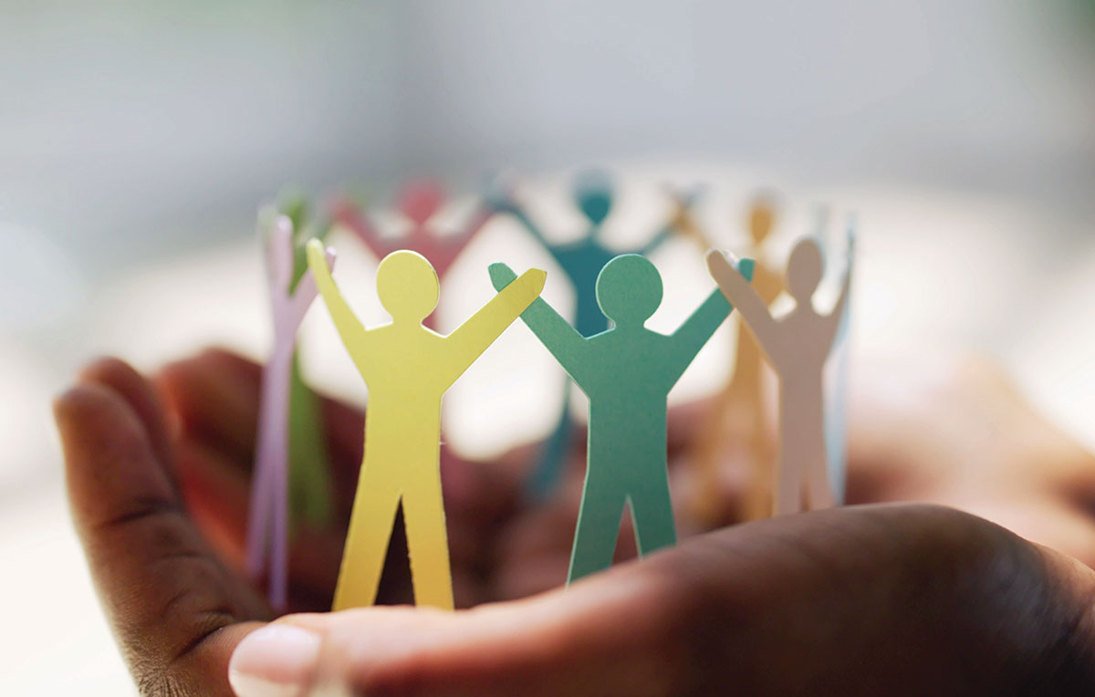 A human hand holding paper dolls arranged in a circle that are multi-colored and holding each other's raised hands