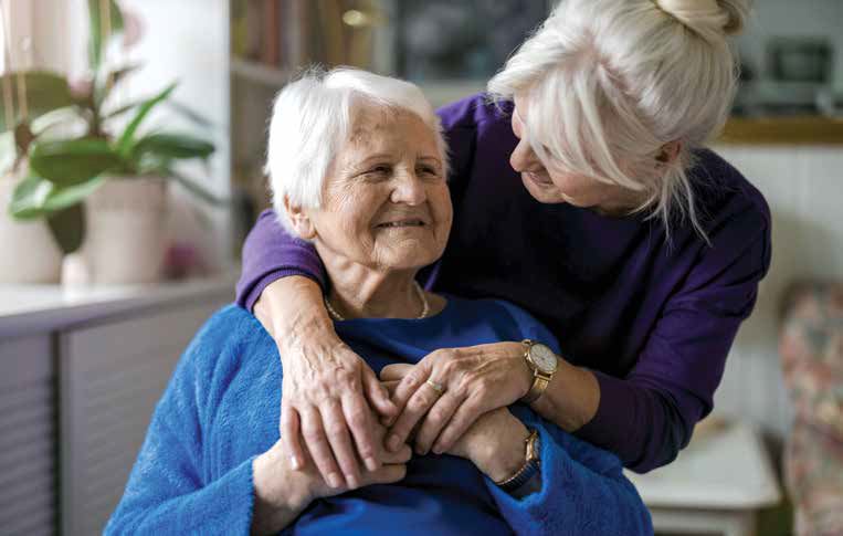 Woman standing and hugging a woman from behind who is sitting; both are smiling.