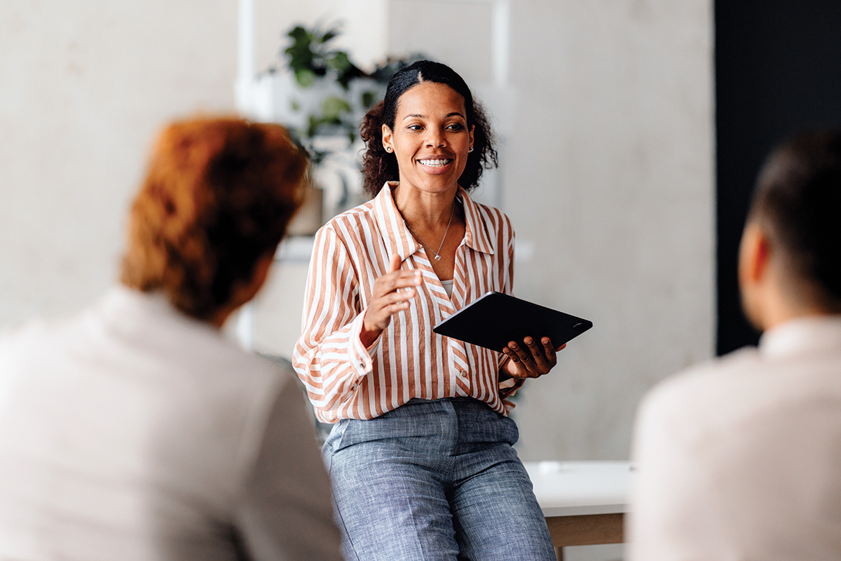 Woman smiling, holding tablet, and sitting on desk, facing two other people and gesturing to them