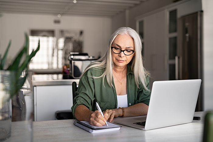 Woman writing at desk at home with open laptop