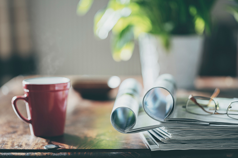 Desk with stack of magazines and a pair of glasses on top alongside a cup of coffee and a plant