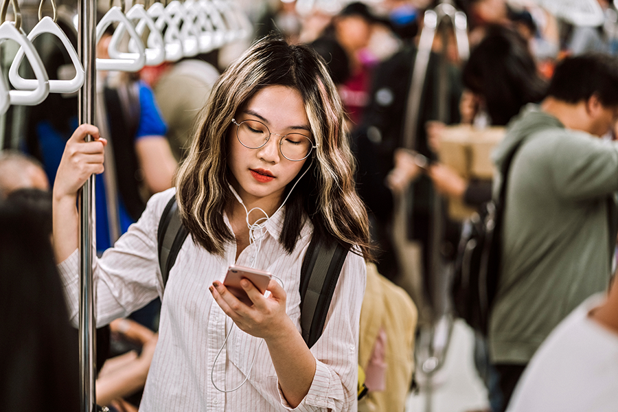 Young female holding pole on subway train with earbuds in her ears listening to podcasts on her mobile phone