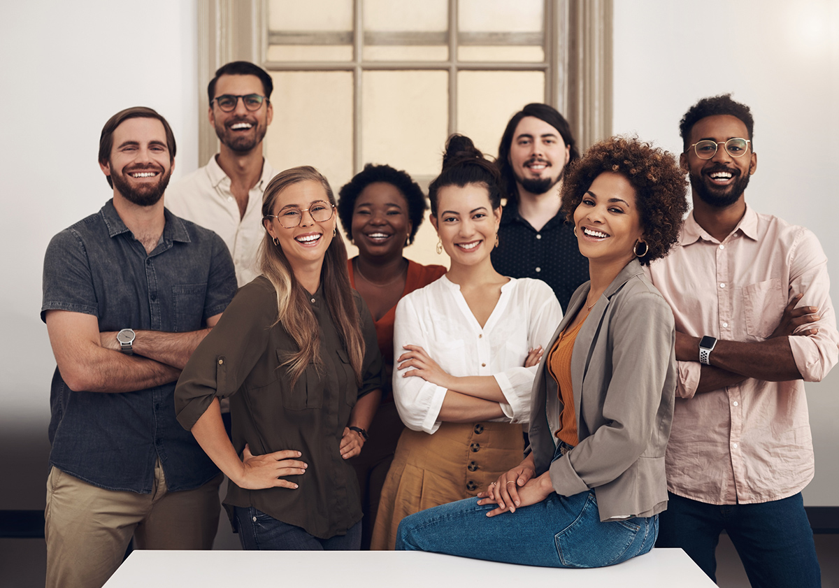 Large diverse group of coworkers standing around table inside office smiling with one woman sitting on table