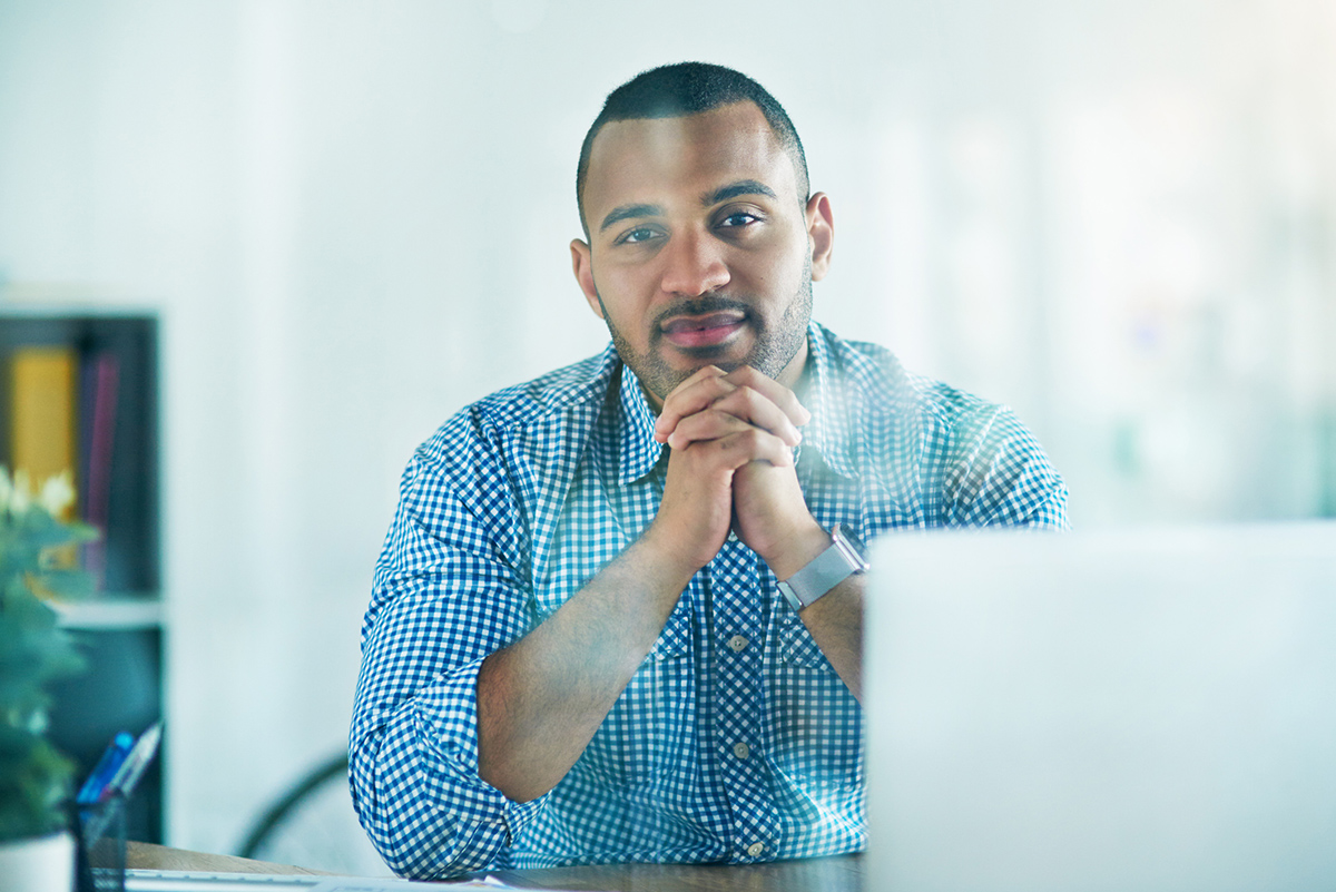 Man with hands clasped under chin sitting at table with open laptop looking at camera