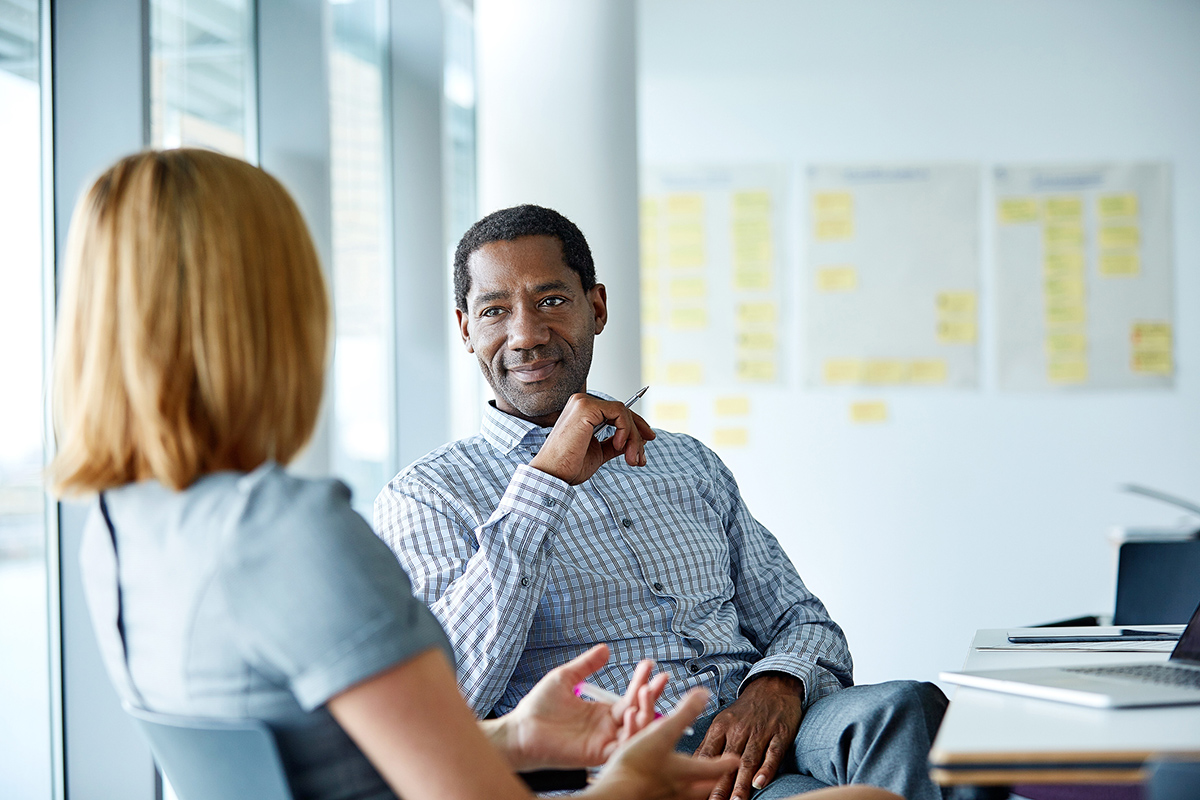 Professional man leaning back in chair listening to female coworker talk inside office