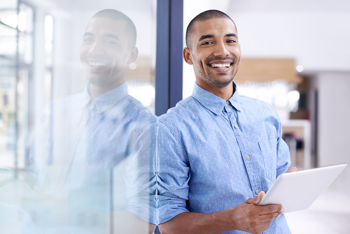 Smiling young professional man in business casual leaning on office wall holding a tablet