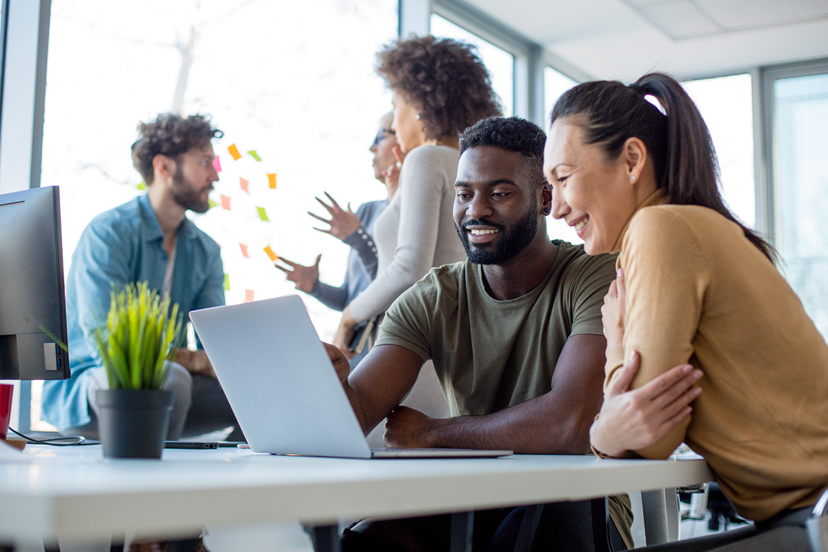 One male and one female sitting next to each other smiling and looking at open laptop with coworkers in the background