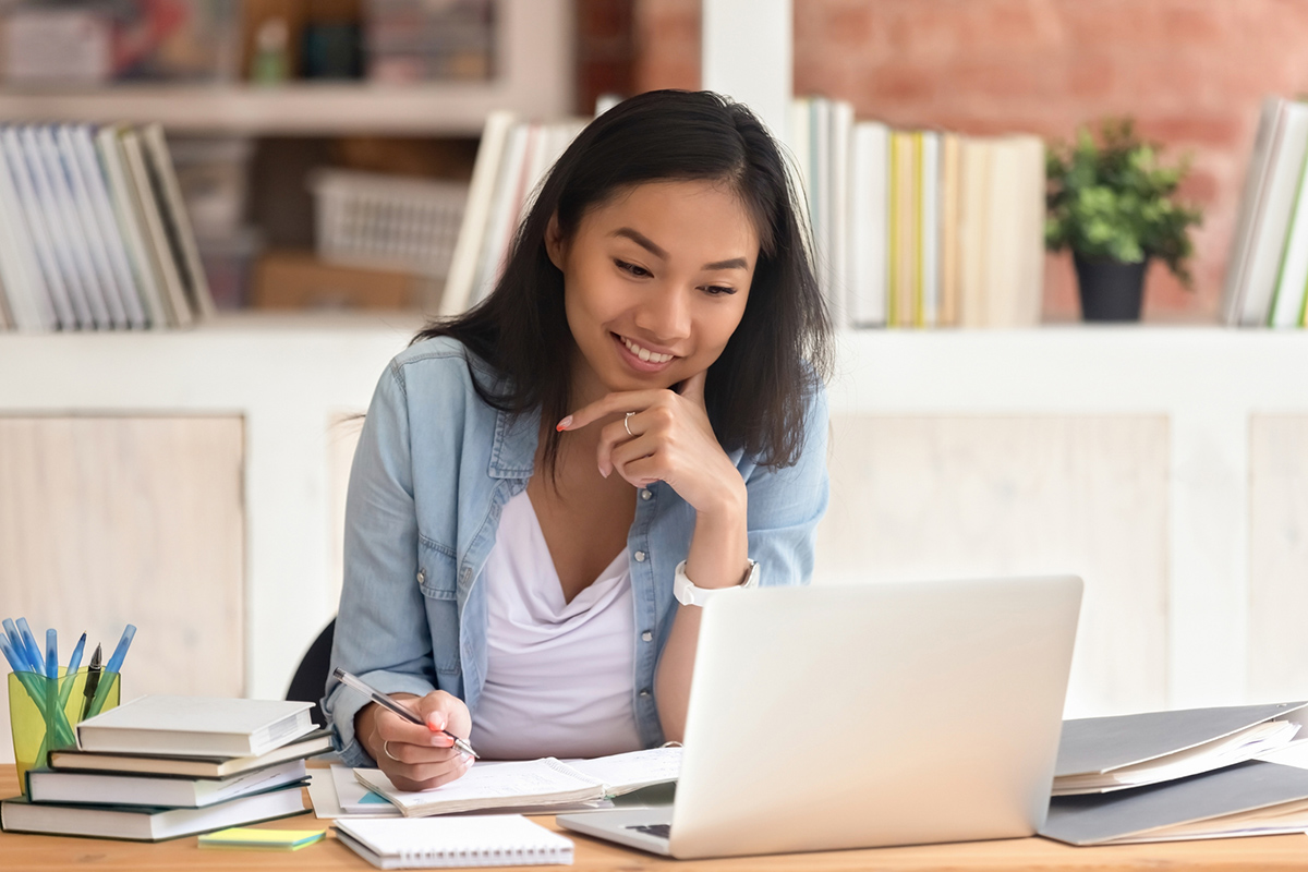 Smiling woman sitting at desk looking at laptop with pen in hand taking research notes