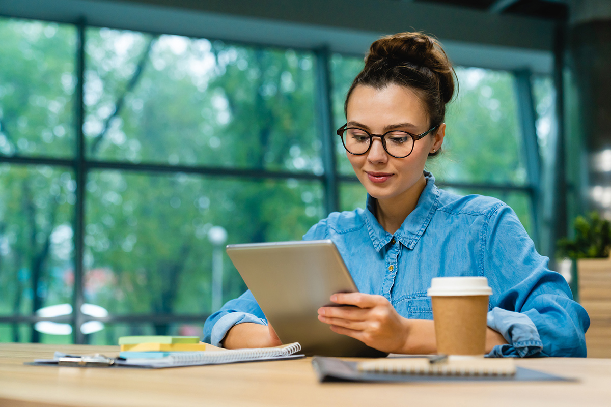 Woman wearing glasses sitting at a table with coffee reading a tablet OT Practice