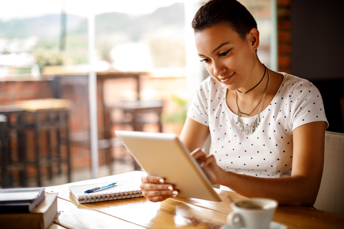 Young woman sitting at cafe table reading learning resources on her tablet