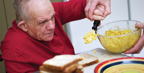 An older adult spoons food onto some bread with some assistance 