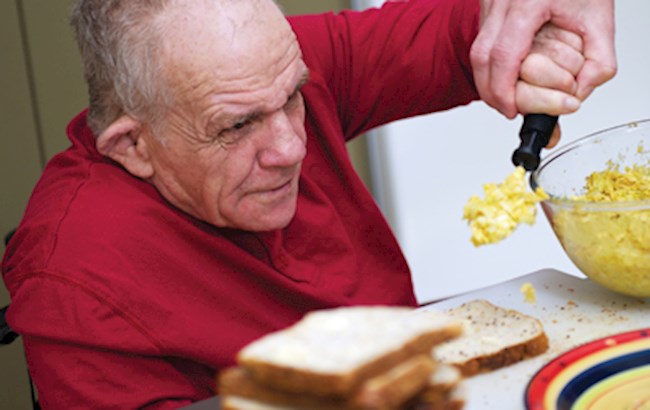 An older adult spoons food onto some bread with some assistance 