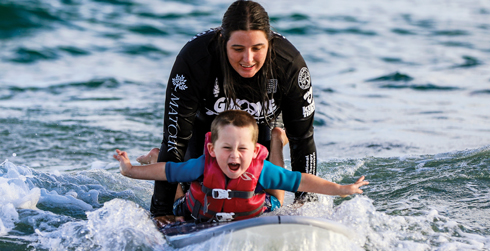 An athlete rides a wave on his stomach, arms outstretched and smiling, with his surf therapist riding behind him.