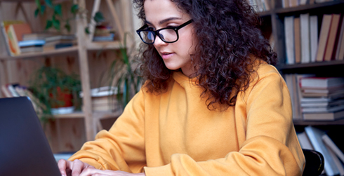 Girl with glasses typing on laptop. Bookcases in the background
