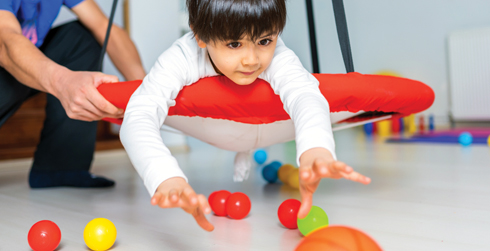 Child laying down on their stomach in a swing while playing with balls on the floor