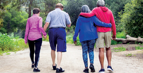 Four adults walking on a path in the woods. The two adults on the left are holding hands, while the other two adults on the right have their arms around each other.