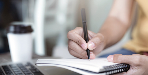 Hand writing with pen in notebook, coffee cup nearby.
